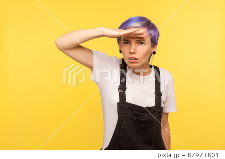 Portrait of wondered young hipster girl with violet short hair in denim overalls holding hand above eyes and looking far away with attentive curious view. isolated on yellow background, studio shot Portrait of wondered young hipster girl with violet short hair in denim overalls holding hand above eyes and looking far away with attentive curious view. isolated on yellow background, studio shot 87973801