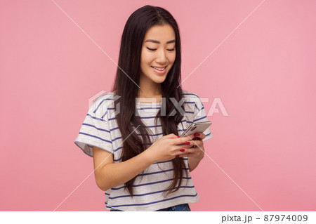 Portrait of happy girl with brunette hair in t-shirt typing message on mobile phone and smiling, reading funny post in social network, chatting online. indoor studio shot isolated on pink background 87974009