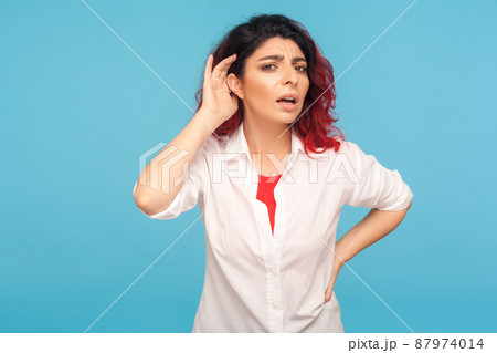 Portrait of nosy hipster woman with fancy red hair in shirt listening attentively holding hand near ear to hear confidential talk, hard to understand. indoor studio shot isolated on blue background 87974014