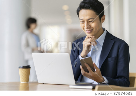 オフィスワーク イメージ スマートフォンを持ってパソコンに向かうビジネスマン オフィスワーク イメージ スマートフォンを持ってパソコンに向かうビジネスマン 87974035