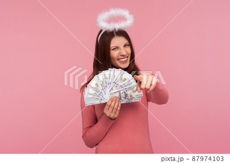 Cheerful brunette woman with halo above her head holding dollar banknotes pointing finger at you with toothy smile, lottery winner, success. Indoor studio shot isolated on pink background Cheerful brunette woman with halo above her head holding dollar banknotes pointing finger at you with toothy smile, lottery winner, success. Indoor studio shot isolated on pink background 87974103
