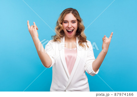 Portrait of crazy funky enthusiastic woman with wavy hair in white jacket showing rock and roll sign and screaming for joy, doing devil horns gesture. indoor studio shot isolated on blue background Portrait of crazy funky enthusiastic woman with wavy hair in white jacket showing rock and roll sign and screaming for joy, doing devil horns gesture. indoor studio shot isolated on blue background 87974166