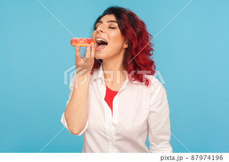 Hungry woman with fancy red hair biting donut with expression of pleasure, eating delicious pastry, can't resist temptation, addicted to sugar, high calorie food. indoor studio shot, blue background 87974196