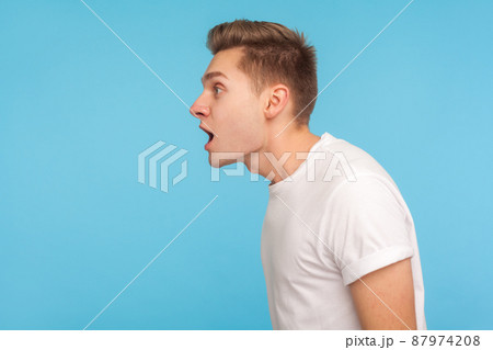 Wow, amazing. Side view of astonished man in casual white t-shirt standing with mouth open in surprise, scared and shocked by crazy unbelievable news. indoor studio shot isolated on blue background 87974208