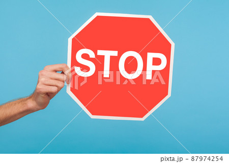 Closeup of male hand holding Stop symbol, showing red traffic sign, warning about problems, concept of ban, denial, prohibited to go, forbidden way. indoor studio shot isolated on blue background Closeup of male hand holding Stop symbol, showing red traffic sign, warning about problems, concept of ban, denial, prohibited to go, forbidden way. indoor studio shot isolated on blue background 87974254