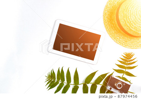 Straw hat with green leaves, tablet and old camera on white background, Summer background. Top view. Sun flare Straw hat with green leaves, tablet and old camera on white background, Summer background. Top view. Sun flare 87974456