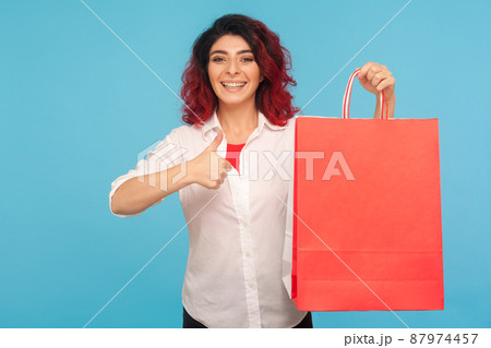 Great shopping. Portrait of excited shopaholic hipster woman with fancy red hair holding packages and showing thumbs up, satisfied with weekend sale. indoor studio shot isolated on blue background Great shopping. Portrait of excited shopaholic hipster woman with fancy red hair holding packages and showing thumbs up, satisfied with weekend sale. indoor studio shot isolated on blue background 87974457