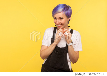 Portrait of sly hipster woman with violet short hair in denim overalls smirking and looking at camera with cunning face, thinking devious tricks and cheats. isolated on yellow background, studio shot 87974467