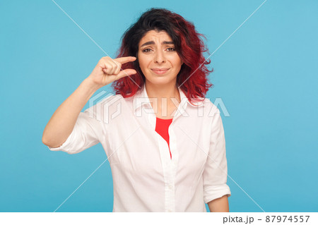 Small size. Portrait of displeased hipster woman with fancy red hair making a little bit gesture and looking with dissatisfied face, showing minimum. indoor studio shot isolated on blue background Small size. Portrait of displeased hipster woman with fancy red hair making a little bit gesture and looking with dissatisfied face, showing minimum. indoor studio shot isolated on blue background 87974557