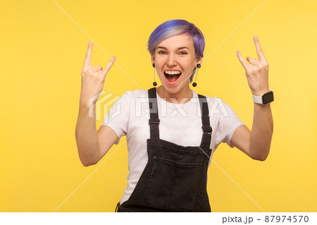 Portrait of delighted funky hipster woman with violet short hair in overalls showing rock and roll gesture, crazy punk sign, devil horns and shouting with happiness. yellow background, studio shot 87974570