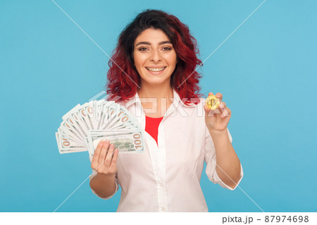 Cryptocurrency exchange. Joyful smiling happy woman with fancy red hair holding bitcoin and dollar banknotes, digital money, electronic commerce. indoor studio shot isolated on blue background 87974698