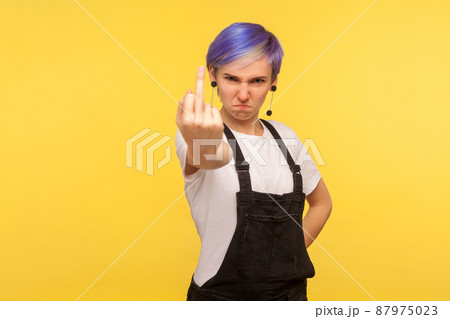 Portrait of crazy angry hipster woman with violet hair in denim overalls showing middle finger, rude fuck off gesture, face full of hatred and aggression. isolated on yellow background, studio shot 87975023