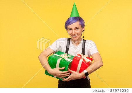 Portrait of happy joyful hipster girl with violet short hair wearing funny party cone and overalls embracing gift boxes, satisfied with presents, enjoying birthday party. yellow background studio shot Portrait of happy joyful hipster girl with violet short hair wearing funny party cone and overalls embracing gift boxes, satisfied with presents, enjoying birthday party. yellow background studio shot 87975056