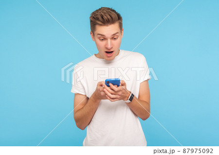 Portrait of amazed astonished man in white t-shirt using cellphone with surprised expression, shocked by message in social network, mobile communication. indoor studio shot isolated on blue background 87975092