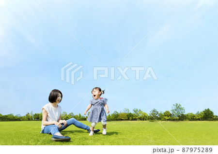 青空を背景に緑の芝生の上で楽しく遊ぶ母と幼い女の子。親子,幸せ,愛情,育児のイメージ	 87975289