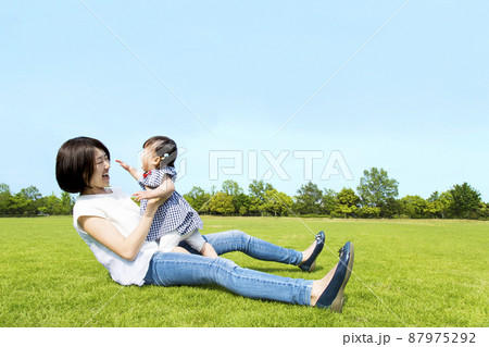 青空を背景に緑の芝生の上で楽しく遊ぶ母と幼い女の子。親子,幸せ,愛情,育児のイメージ	 87975292