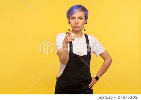 Hey you. Portrait of hipster woman with violet short hair in denim overalls pointing at camera and looking strict rigid, making choice with serious face, selecting you. yellow background, studio shot 87975308