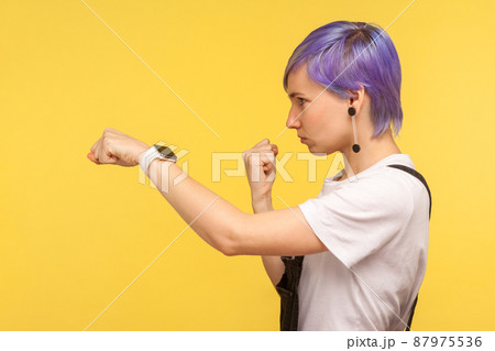 Side view of determined young hipster woman with violet dyed short hair in denim overalls punching with fist, showing boxing gesture, looking confident purposeful. yellow background, studio shot Side view of determined young hipster woman with violet dyed short hair in denim overalls punching with fist, showing boxing gesture, looking confident purposeful. yellow background, studio shot 87975536