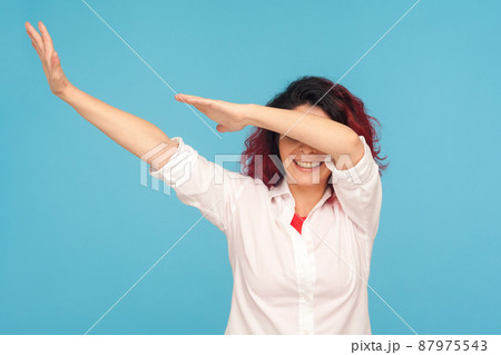Happy cheerful woman in white shirt showing dab dance move, celebrating winning with famous internet meme of success, performing dabbing youth trends. indoor studio shot isolated on blue background 87975543
