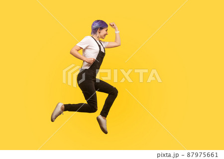 Side view full length, enthusiastic energetic hipster girl with violet hair in overalls jumping or flying in air, running quickly fast, hurrying for sales. isolated on yellow background, studio shot 87975661
