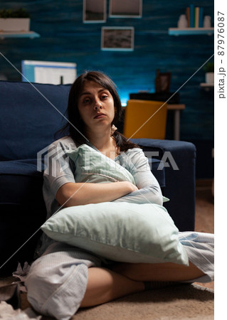 Portrait of depressed woman with mental health problems sitting on floor and hugging pillow in living room. Anxious sad person dealing with chronic disease difficulties and depression at home. Portrait of depressed woman with mental health problems sitting on floor and hugging pillow in living room. Anxious sad person dealing with chronic disease difficulties and depression at home. 87976087