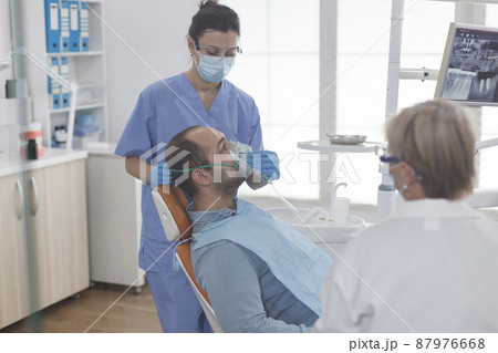 Man patient sitting on dental chair during stomatology examination while dentist assistant putting oxygen mask before teeth surgery in stomatological office room. Medical team wearing face mask 87976668