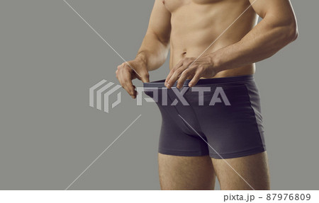 Handsome young male looks inside underpants at his private area. Shirtless attractive muscular man in underwear briefs looking at his penis standing isolated on grey background. Genital health concept 87976809