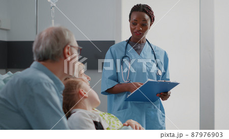 African american nurse talking to elder patient and family in hospital ward. Medical assistant explaining diagnosis to senior woman and visitors. Healthcare specialist discussing treatment. African american nurse talking to elder patient and family in hospital ward. Medical assistant explaining diagnosis to senior woman and visitors. Healthcare specialist discussing treatment. 87976903