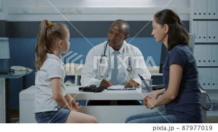 Doctor talking to patients and taking notes on computer to prepare checkup document with prescription medicine. Medic having conversation with little girl and adult in medical cabinet. 87977019