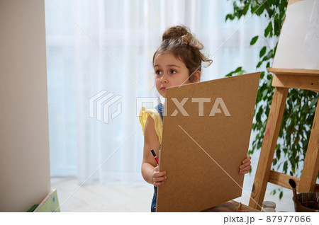 Adorable little preschool girl stands at a wooden easel and looks away while holding a pencil and a canvas. Drawing, painting and art creativity concept. Education and kids entertainment concept 87977066