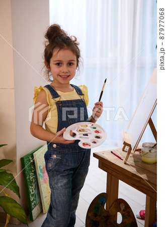Charming little girl, talent artist, adorable child holding a paintbrush and palette, standing near a wooden easel while enjoying art class. Kids entertainment, hobby, education, creativity 87977068