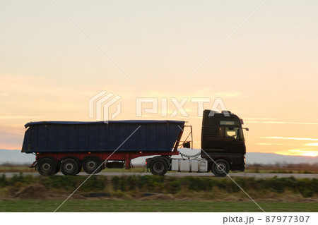 Semi-truck with tipping cargo trailer transporting sand from quarry driving on highway hauling goods in evening. Delivery transportation and logistics concept 87977307