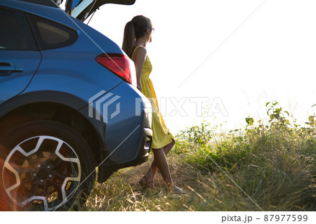 Happy young woman driver in yellow dress enjoying warm summer evening standing beside her car. Travelling and vacation concept. 87977599