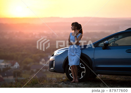 Happy young woman driver in blue dress enjoying warm summer evening standing beside her car. Travelling and vacation concept. Happy young woman driver in blue dress enjoying warm summer evening standing beside her car. Travelling and vacation concept. 87977600