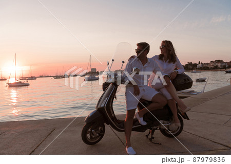 The couple enjoys a vacation while sitting on a scooter by the sea and enjoying the beautiful sunrise. Selective focus  87979836