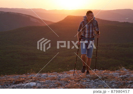 Young hiker with backpack and trekking poles walking along a crimea mountain path at sunset with a smile. High quality photo. Young hiker with backpack and trekking poles walking along a crimea mountain path at sunset with a smile. High quality photo. 87980457