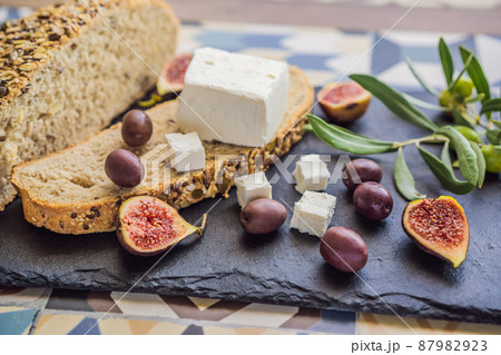 Green and black olives with loaf of fresh bread, feta cheese and young olives branch on olive wood chopping board over dark background Green and black olives with loaf of fresh bread, feta cheese and young olives branch on olive wood chopping board over dark background 87982923