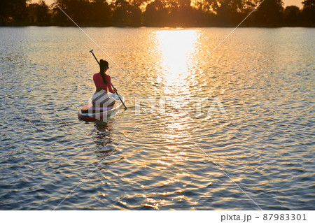 Active caucasian woman dressed in colorful sport clothes swimming with paddle on sup board during evening time. Amazing summer sunset over lake. Water sport concept. Active caucasian woman dressed in colorful sport clothes swimming with paddle on sup board during evening time. Amazing summer sunset over lake. Water sport concept. 87983301