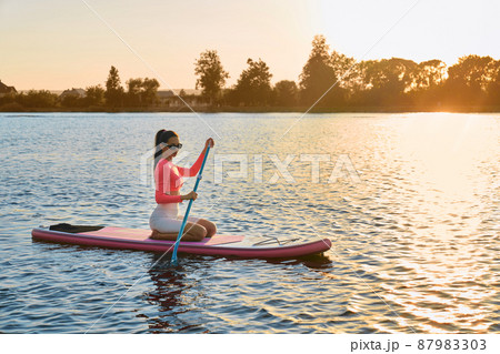 Young woman with athletic body shape swimming on sup board with long paddle in hands. Active female rowing on fresh air with amazing sunset on background. Young woman with athletic body shape swimming on sup board with long paddle in hands. Active female rowing on fresh air with amazing sunset on background. 87983303