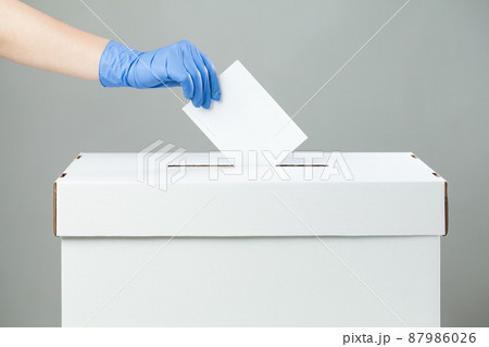 Caucasian female hand wearing blue protective latex rubber glove placing ballot paper in vote box Caucasian female hand wearing blue protective latex rubber glove placing ballot paper in vote box 87986026