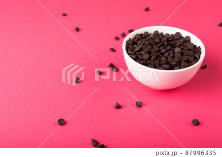 High angle view of fresh chocolate chips full of white bowl against pink background 87996335