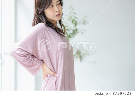 部屋着のまま腰に手を当てる体調不良の女性 87997523