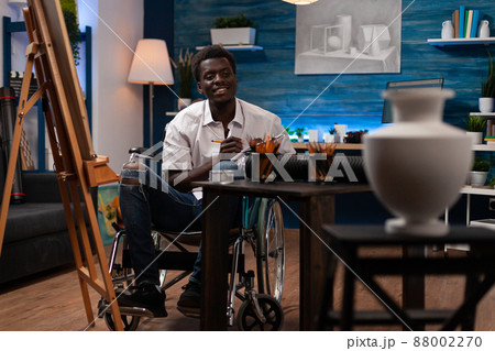 Young illustrator in wheelchair looking at vase for inspiration drawing sketch on notebook working at creative masterpiece in art studio. Artist man with disability sketching project illustration 88002270