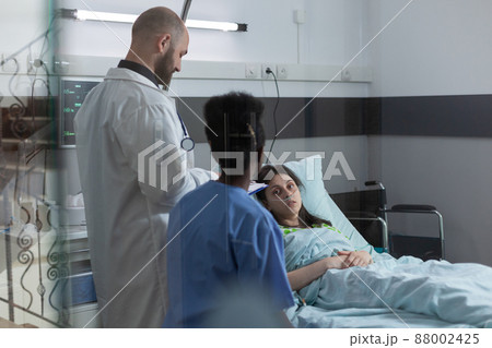 Medical doctor and nurse doing consult in private ward to recovering woman in hospital bed after surgical intervention. Patient with nasal cannula looking at health care professionals. 88002425