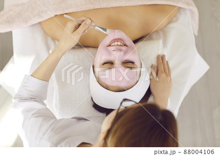 Cosmetology and medical services office. Top view of unknown female beautician with brush applies pink mask on face of satisfied client. Concept of cosmetic procedures in a medical center. 88004106