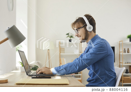 Young happy man in headphones typing on laptop and using internet web online services. Man relaxing and playing games or working remotely at home using modern technology and communication devices. 88004116
