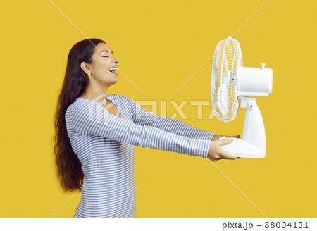 Device for cooling air. Satisfied woman suffering from heat enjoys cold air from electric fan on yellow background. Beautiful smiling woman holding fan in front of her. Hot weather concept. Device for cooling air. Satisfied woman suffering from heat enjoys cold air from electric fan on yellow background. Beautiful smiling woman holding fan in front of her. Hot weather concept. 88004131