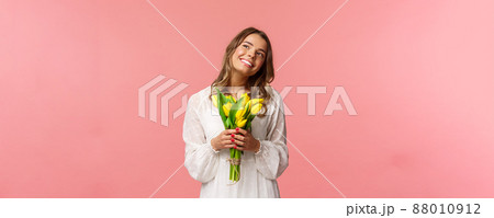 Holidays, beauty and spring concept. Portrait of dreamy, happy blond girl feeling romantic daydreaming about her girlfriend, holding yellow tulips, wear white dress, standing pink background 88010912