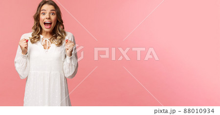 Portrait of surprised lucky hopeful, blond girl winning, hear great news and chanting, gasping open mouth smiling joyful, jumping from happiness and joy, standing pink background Portrait of surprised lucky hopeful, blond girl winning, hear great news and chanting, gasping open mouth smiling joyful, jumping from happiness and joy, standing pink background 88010934