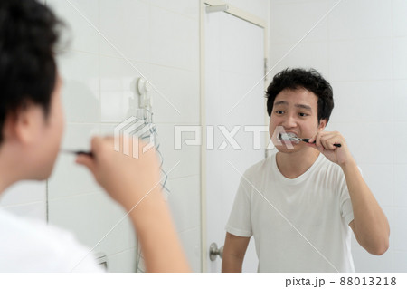 Happy Asian young man in white t-shirt brushing a teeth in bathroom in the morning. A man preparing and checking his face and body before go to work. Routine activity in daily life. Happy Asian young man in white t-shirt brushing a teeth in bathroom in the morning. A man preparing and checking his face and body before go to work. Routine activity in daily life. 88013218